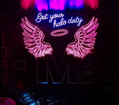 Pink and blue neon sign in a dark lounge: glowing angel wings and halo with cursive text 'Get your halo dirty', colorful reflections on wall and floor