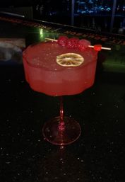 Bright pink raspberry cocktail in a scalloped coupe glass on a dark bar counter, garnished with skewered raspberries and a dried lemon wheel — vibrant bar nightlife drink.