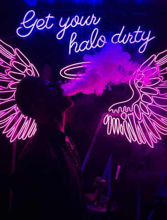 Silhouette of a person exhaling purple vapor in front of pink and blue neon wings and halo with the sign Get your halo dirty, neon-lit hookah vibe