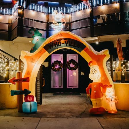 Nighttime holiday storefront entrance with inflatable "Welcome to Who-Ville" arch, Grinch characters, "Grinch Lane" sign, wrapped gift, wreath-adorned double doors and twinkling string lights.