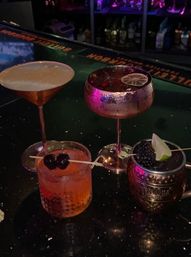 Four colorful cocktails on a dim bar counter with purple ambient lighting: a frothy drink in a copper coupe, a metallic goblet topped with cranberries and a lime wheel, an old-fashioned garnished with two cherries on a skewer, and a copper mule mug with a blackberry and lime wedge.