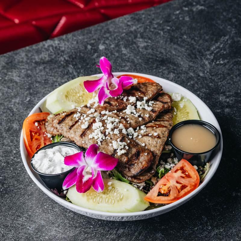Grilled steak salad bowl topped with crumbled feta, cucumber slices, tomato wedges, two dipping sauces and a bright pink orchid garnish on a dark stone tabletop