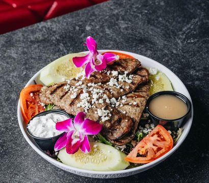 Grilled steak salad bowl topped with crumbled feta, cucumber slices, tomato wedges, two dipping sauces and a bright pink orchid garnish on a dark stone tabletop