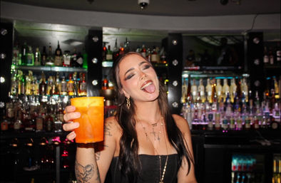 Playful person at a dimly lit bar holding a bright orange cocktail to the camera, tongue out, septum piercing and tattoos, long dark hair and black strapless top with backlit liquor bottles on shelves behind