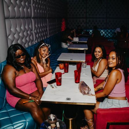 Four friends posing at a booth in a dim urban lounge — tufted white walls, teal banquette seating, marble tables and red drink cups for a lively nightlife girls' night out.