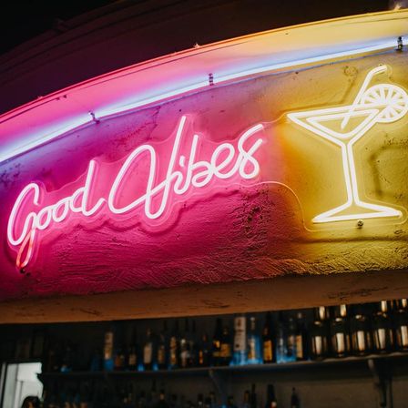 Pink and yellow neon sign reading 'Good Vibes' with a martini glass glowing above shelves of liquor bottles in a bar interior