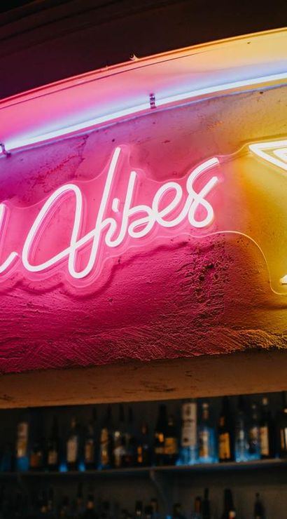 Pink and yellow neon sign reading 'Good Vibes' with a martini glass glowing above shelves of liquor bottles in a bar interior