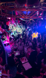 Overhead view of a crowded nightclub dance floor beneath a large disco ball, colorful pink, purple and blue lights illuminating people dancing and mingling around small tables with red cups.