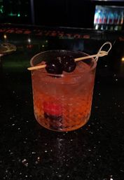 Amber old-fashioned cocktail in a cut-glass rocks glass with crushed ice and dark cherries on a bamboo pick, sitting on a glossy black bar counter.