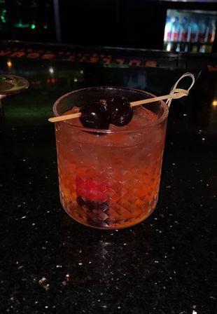 Amber old-fashioned cocktail in a cut-glass rocks glass with crushed ice and dark cherries on a bamboo pick, sitting on a glossy black bar counter.