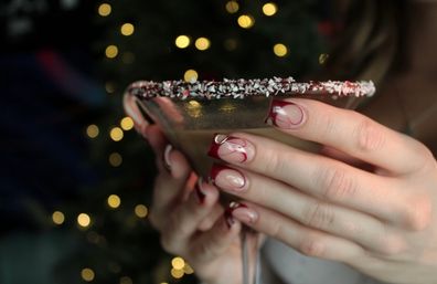 Hands with red-swirled French manicure holding a peppermint-rimmed cocktail glass against warm holiday bokeh lights