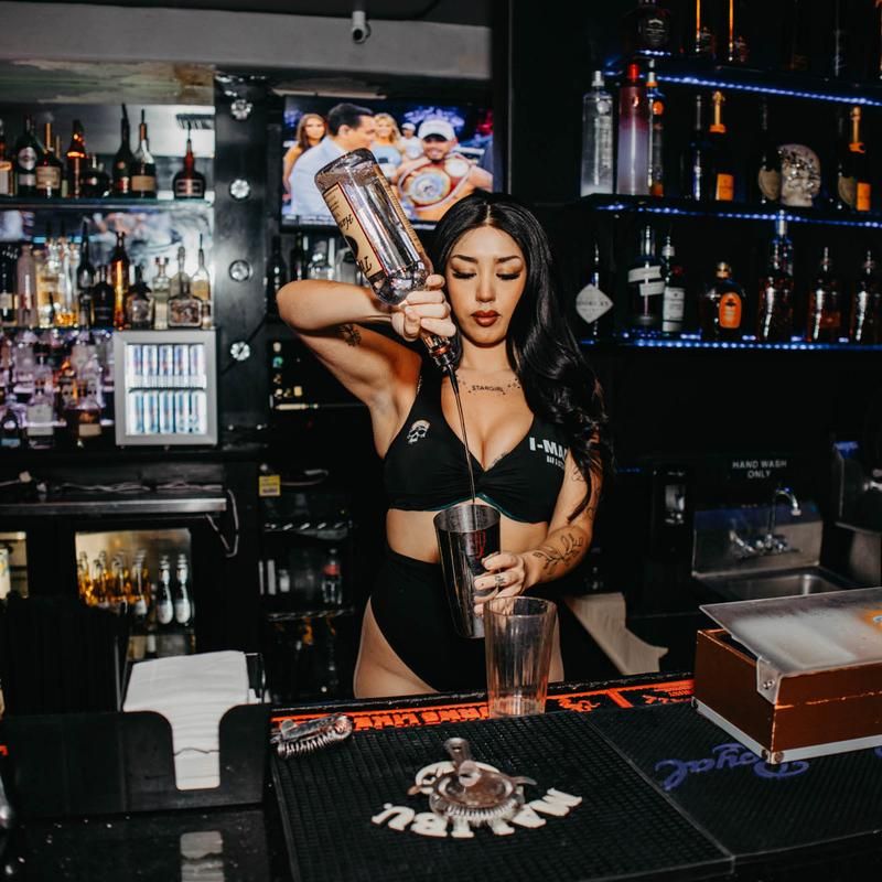 Bartender in black outfit performing a high pour into a cocktail shaker at a neon-lit nightclub bar with shelves of spirits and a TV in the background