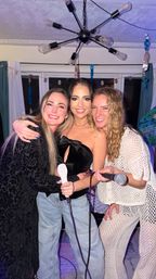 Three friends smiling and hugging while holding microphones at a lively home karaoke party in a living room, under a modern sputnik chandelier with streamers and curtains in the background.