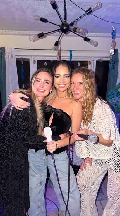 Three friends smiling and hugging while holding microphones at a lively home karaoke party in a living room, under a modern sputnik chandelier with streamers and curtains in the background.