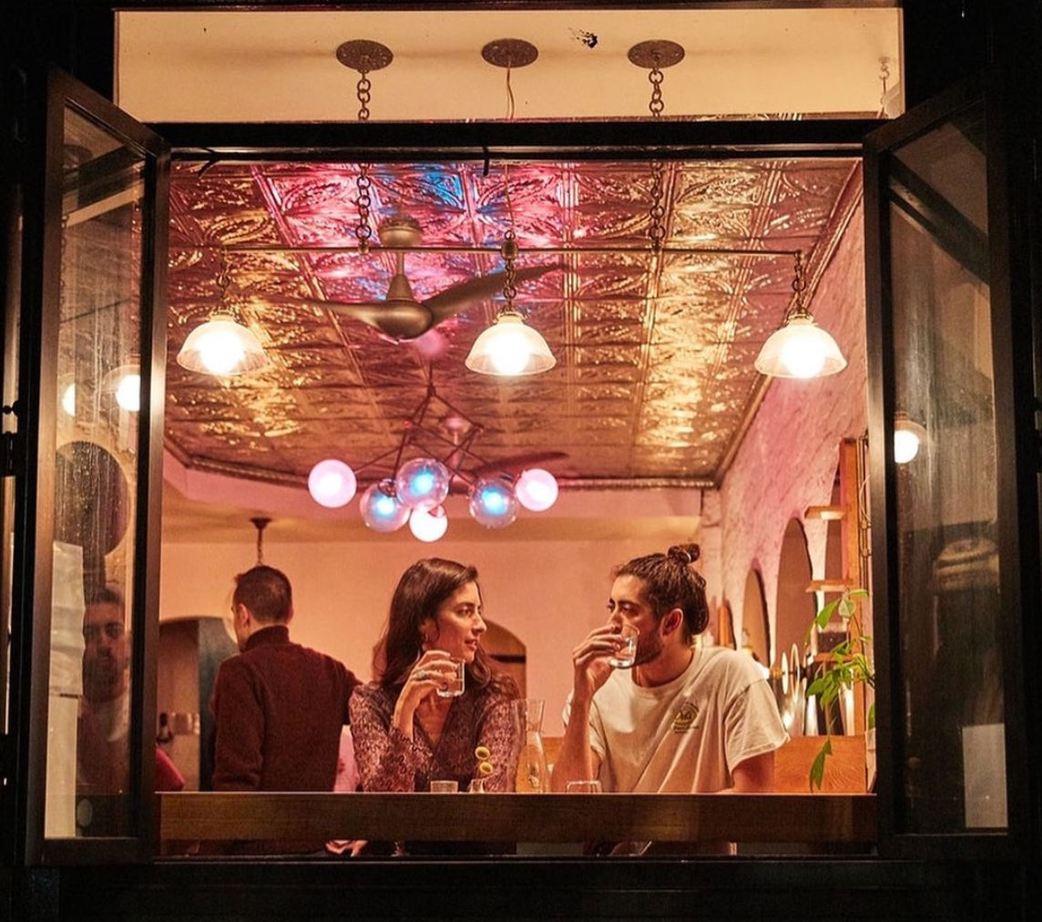 Two people sip drinks at an open-window counter in a cozy urban café at night, warm pendant lights and a colorful globe chandelier glowing off a decorative tin ceiling.