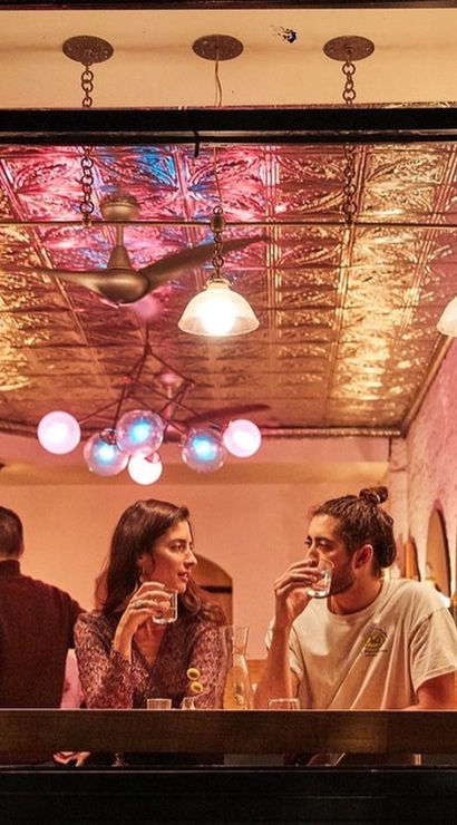 Two people sip drinks at an open-window counter in a cozy urban café at night, warm pendant lights and a colorful globe chandelier glowing off a decorative tin ceiling.