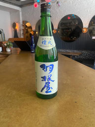 Green glass bottle of Junmai Ginjo sake with bold blue Japanese calligraphy on the label, placed on a wooden bar counter in a cozy modern bar with round mirrors and warm ambient lighting.