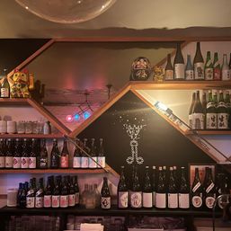 Cozy Japanese-style bar interior with hexagonal wooden shelves filled with rows of sake bottles, ceramic cups, a golden maneki-neko and daruma doll, colorful pendant lights and a chalkboard-style illustration.
