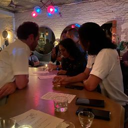 Group of people leaning at a wooden bar in a cozy, dimly lit cocktail bar interior with exposed white brick, colorful red and blue pendant lights, menus and small drink glasses on the counter.