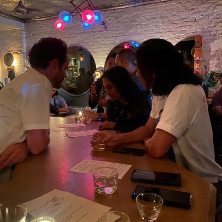 Group of people leaning at a wooden bar in a cozy, dimly lit cocktail bar interior with exposed white brick, colorful red and blue pendant lights, menus and small drink glasses on the counter.