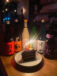 Slice of layered chocolate cake with five lit birthday candles on a white plate, surrounded by six colorful Japanese sake bottles on a dimly lit bar counter with a warm glow