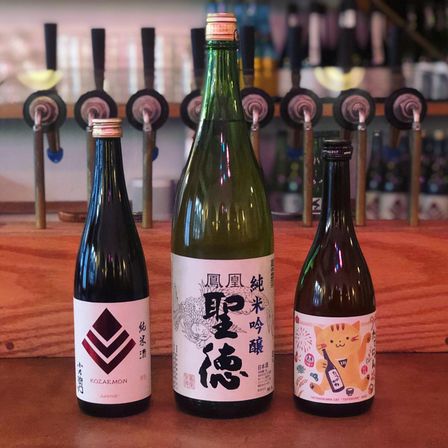 Three Japanese sake bottles on a wooden bar counter with draft taps blurred in the background — a large green bottle centered between two smaller bottles with stylized labels.