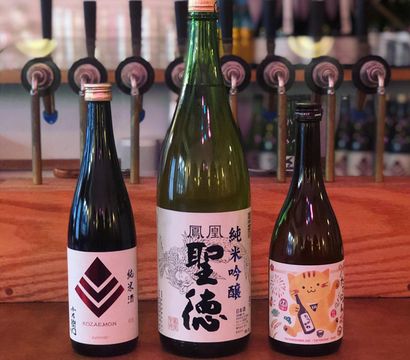 Three Japanese sake bottles on a wooden bar counter with draft taps blurred in the background — a large green bottle centered between two smaller bottles with stylized labels.