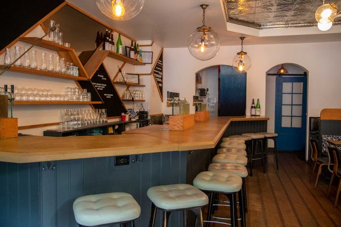 Cozy modern neighborhood bar interior with an L-shaped wooden counter, tufted bar stools, geometric shelves of glassware and bottles, and warm globe pendant lights.