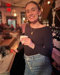 Beaming woman in a black top and button‑fly denim skirt holding a cup at a cozy, dimly lit bar with hanging globe lights and wine bottles on shelves.