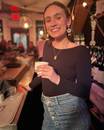 Beaming woman in a black top and button‑fly denim skirt holding a cup at a cozy, dimly lit bar with hanging globe lights and wine bottles on shelves.