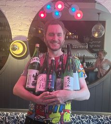 Person smiling and holding multiple large Japanese sake bottles inside a cozy bar, wearing a colorful graphic shirt under pink and blue pendant lights with a round mirror and bartender in the background
