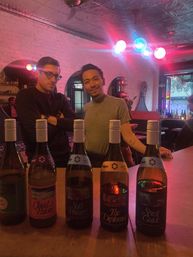 Two people posing behind a bar counter lined with five wine bottles, colorful globe pendant lights casting pink and blue glow, exposed brick walls and tin ceiling in a cozy urban wine bar.