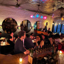 Urban cocktail bar interior at night — cozy, candlelit lounge with exposed white brick, three round mirrors, colorful globe pendant lights and patrons chatting at a busy bar and tables.