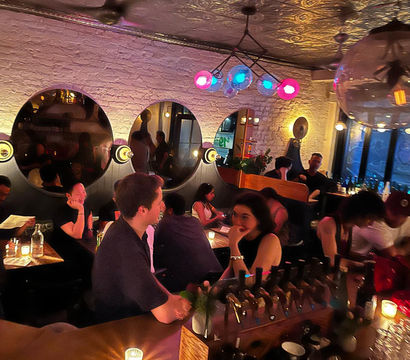 Urban cocktail bar interior at night — cozy, candlelit lounge with exposed white brick, three round mirrors, colorful globe pendant lights and patrons chatting at a busy bar and tables.
