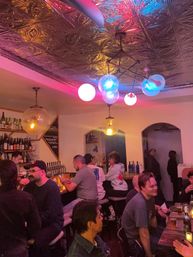 Cozy neighborhood bar interior with ornate tin ceiling and colorful globe pendant lights, patrons seated at the bar and tables, shelves of bottles and beer taps creating a lively evening vibe