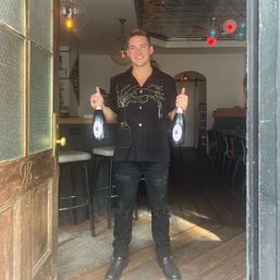 Smiling person in a bar doorway holding two bottles of sparkling wine, wearing a black embroidered shirt and ripped black jeans, ready to celebrate.