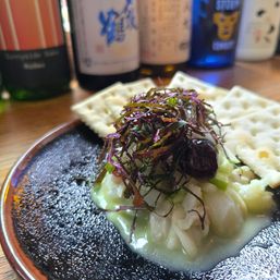 Seafood appetizer of diced white fish in a pale green sauce, topped with shredded purple herbs and a small dark garnish, served with crackers on a black ceramic plate and blurred bottles in the background.