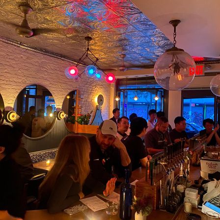 Cozy urban bar interior at dusk — patrons seated along a wooden bar with multiple draft taps, exposed white brick and round mirrors, ornate tin ceiling and colorful globe pendant lights casting red and blue light, city street visible through front windows.