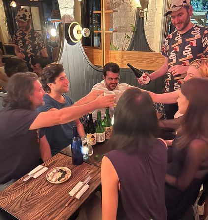 Group of friends clinking ceramic cups while a server pours sake at a cozy, dimly lit urban izakaya-style restaurant — wooden table with sake bottles, small plates and chopsticks, lively nighttime atmosphere.