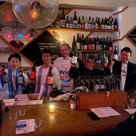 Candlelit Japanese izakaya scene: five people behind a wooden bar holding colorful sake bottles, rows of bottles on shelves, draft taps and a cozy tasting event vibe.