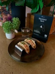 Three mini seafood tacos garnished with dill in a stainless taco holder on a dark plate, beside a glass of water, potted greenery and a tabletop menu card on a restaurant counter.