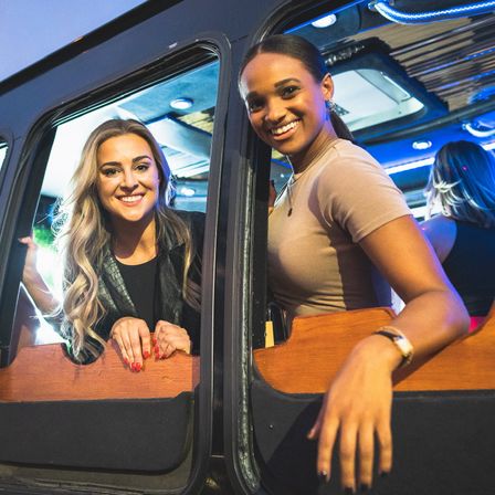 Two smiling women leaning out of a party bus window on a night out, colorful blue interior lights and a festive city nightlife vibe