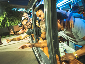Friends leaning out of a party bus at night, laughing, pointing and holding drinks during a lively city nightlife outing