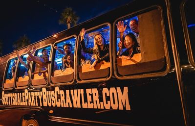 Happy group waving from windows of a nighttime Scottsdale party bus with colorful interior lights and palm trees, lively nightlife scene.