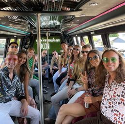 Smiling group of friends in a party bus limo interior, wearing retro-inspired outfits and sunglasses, holding drinks around a center pole under a neon sign reading "you're such an ICON"