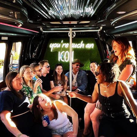 Lively group of friends laughing and drinking inside a party bus, neon sign reading "you're such an ICON" above a center pole