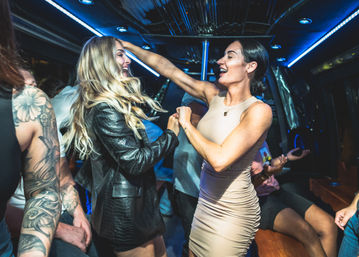 Two friends laughing and dancing inside a neon-lit party bus—blonde in a black leather jacket and a woman in a beige dress, surrounded by seated companions and tattooed arms.