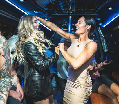 Two friends laughing and dancing inside a neon-lit party bus—blonde in a black leather jacket and a woman in a beige dress, surrounded by seated companions and tattooed arms.