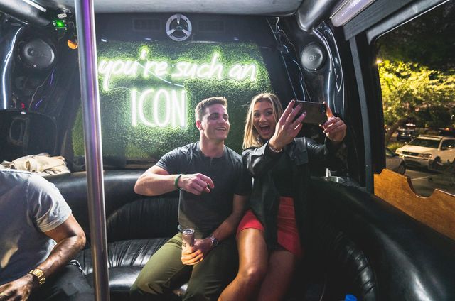 Two friends laughing and taking a selfie inside a nighttime party bus with leather seats, drinks, and a neon sign that reads “you’re such an ICON”