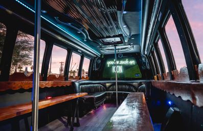 Luxury party bus interior with black leather wrap-around seating, wooden benches, center dance pole, blue LED ceiling lights and a neon sign reading you're such an ICON, pink sunset visible through the windows.